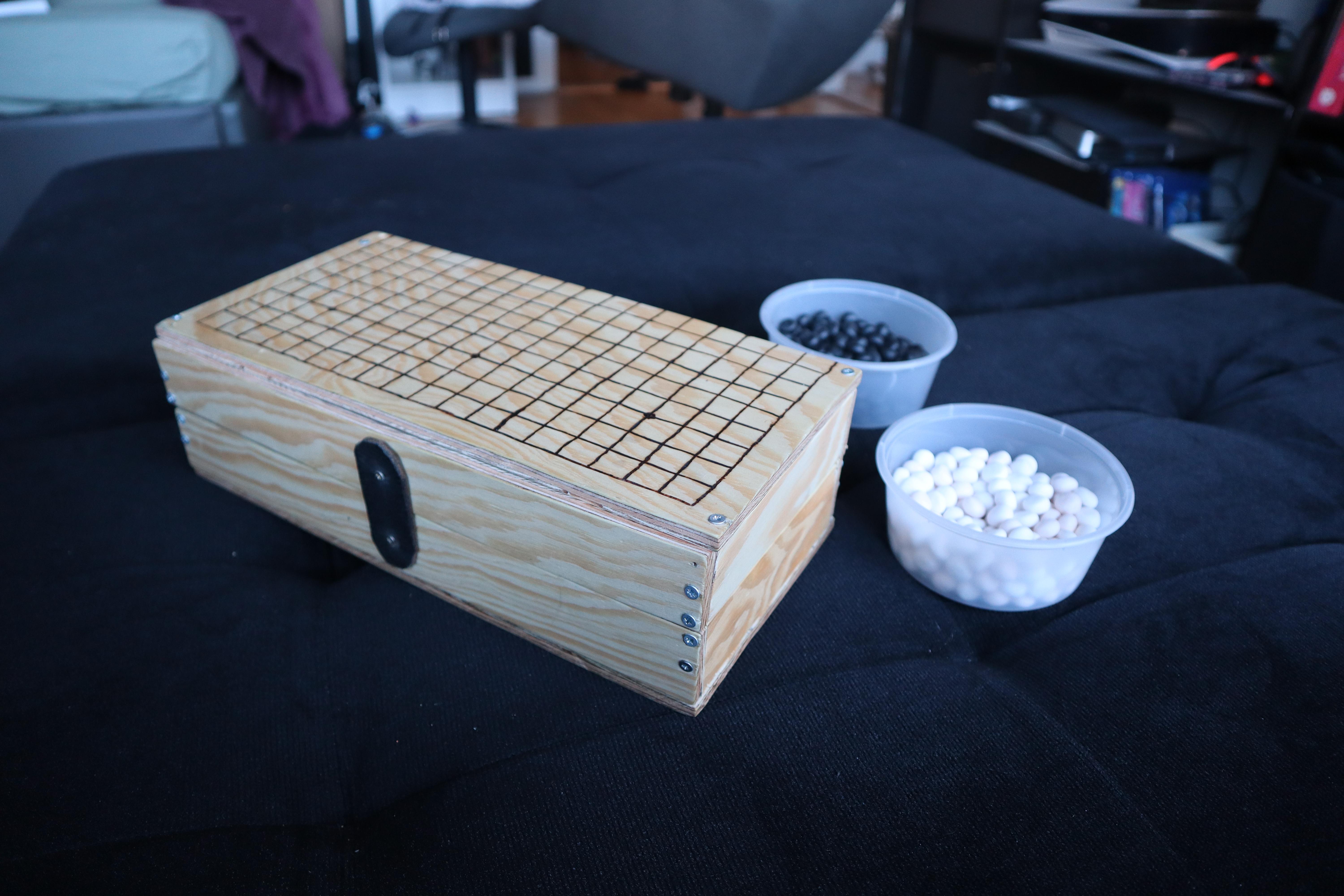 A wooden go board made of plywood and roughly screwed together with burned lines and a leather strap clasp, folded into a box. Beside are two plastic bowls filled with dozens of little round stones made of polymer clay.
