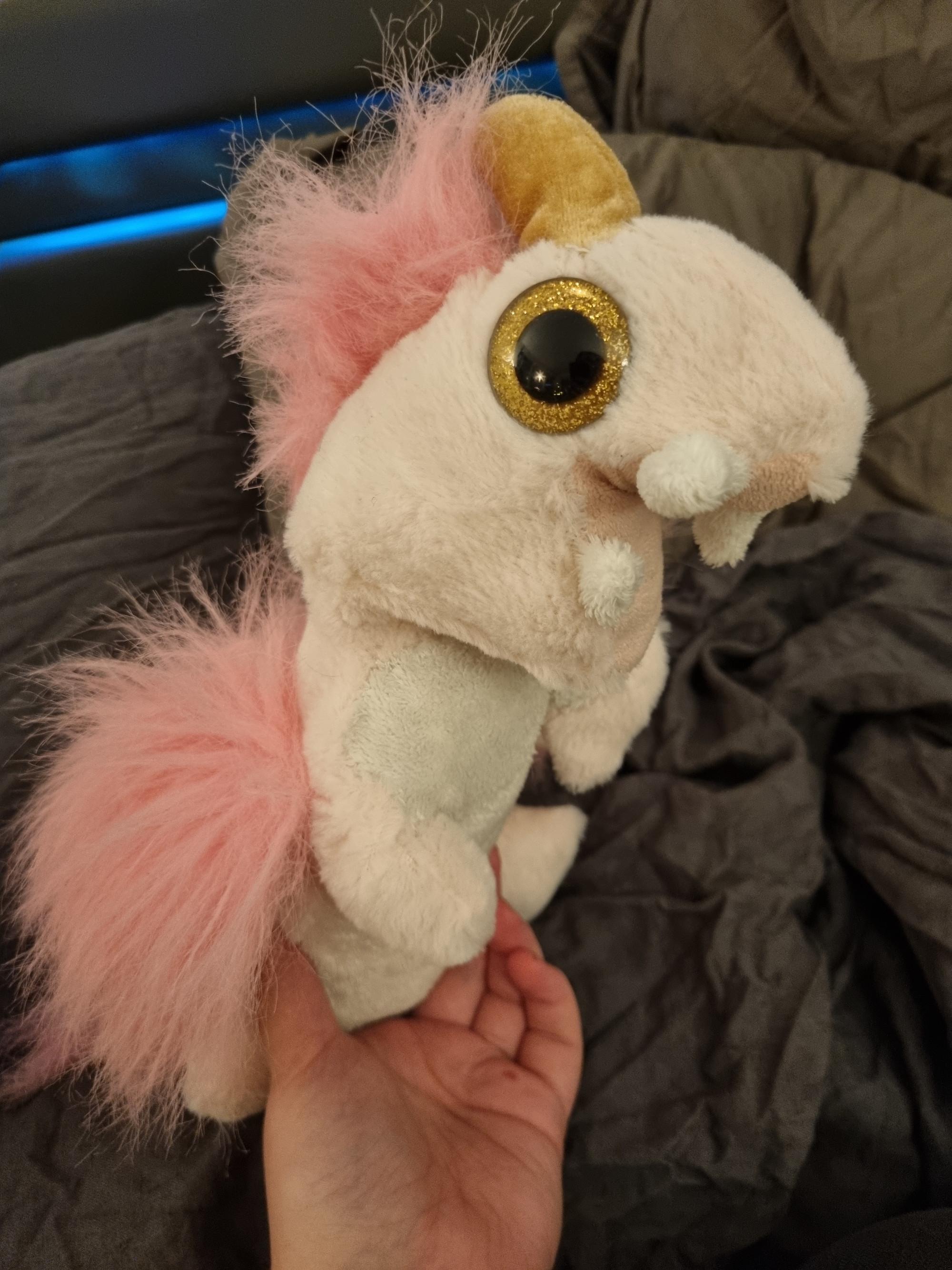 A pink and white plush creature with white body, pink wings, four tusky fangs, a giant golden eye, and a golden horn pointing from it's head.