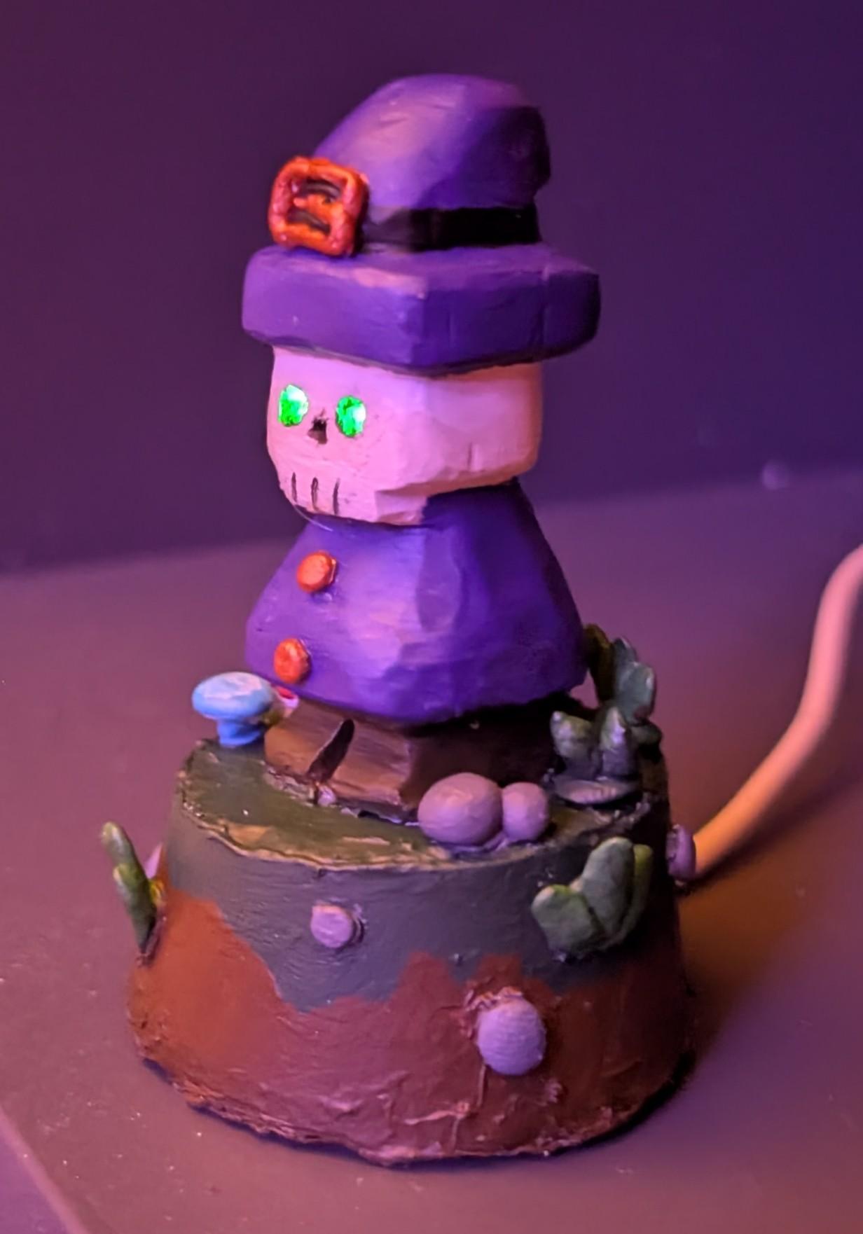 A lil skeleton guy in a purple coat and hat with a copper buckle on the hat and copper buttons on the coat. His eyes glow green and he is on a little base painted with grass and dirt surrounded by stones, leafy grasses, and mushrooms.