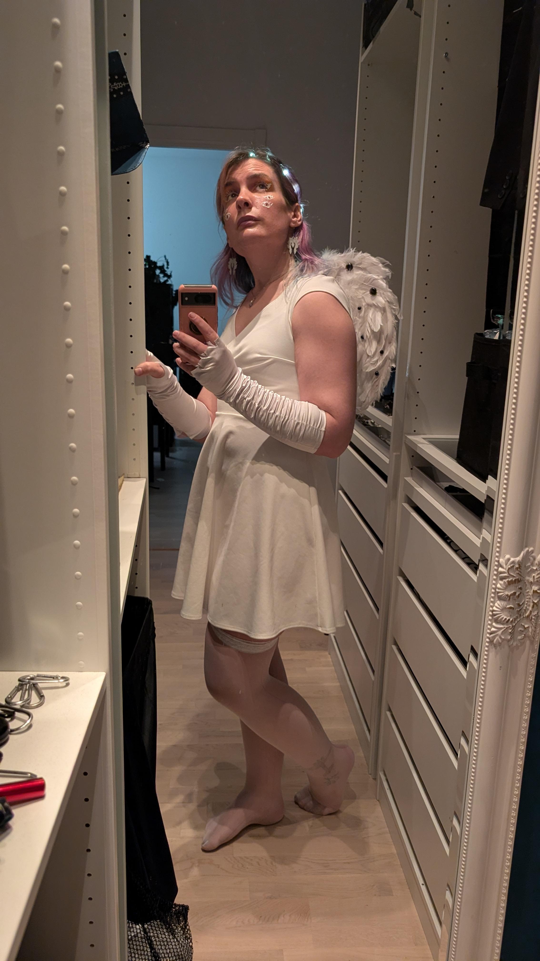 Ash in angel outfit lit from above, white feathered wings covered in eyes on display