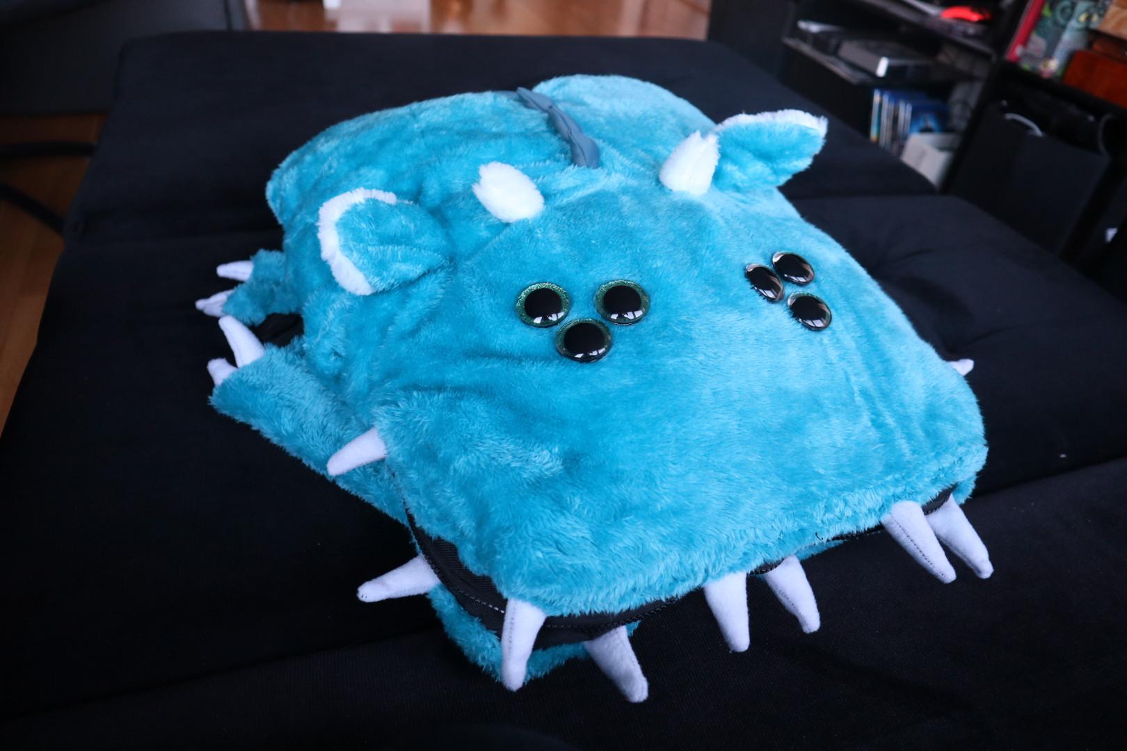 Bright blue cyan plush monster backpack closed with mouth zipper closed, its two sets of triple eyes staring off into space