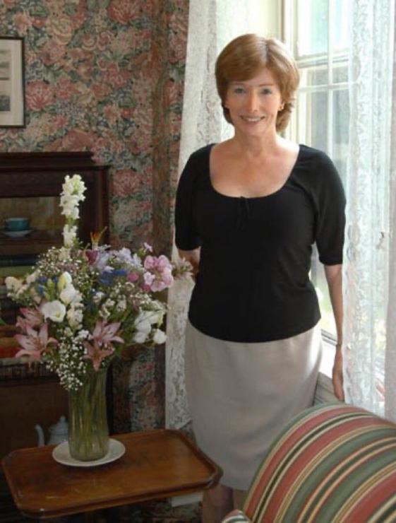 A woman with red hair in a black shirt and light grey skirt, smiling, standing in front of a curtained window and floral wallpaper, behind a striped cushioned chair or sofa and dark wooden table with vase and bouquet of white, blue and pink flowers.