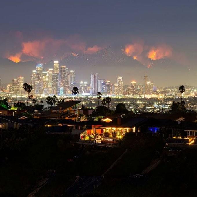 A photo of downtown LA with its skyscrapers illuminate at night, and behind them the hills are ablaze with the Bridge Fire