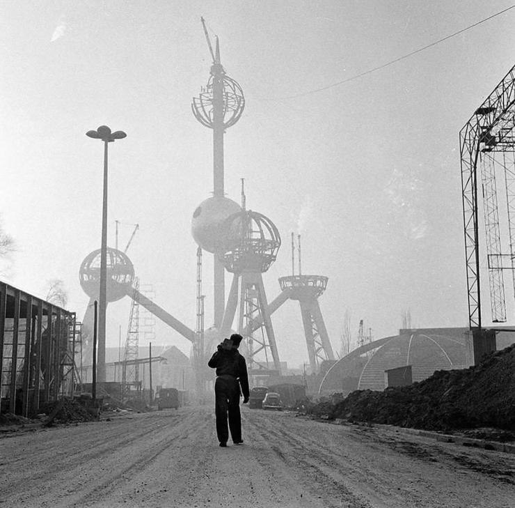 Black and White photo of a construction worker’s silhouette in front of the under-construction Atomium in Brussels (large balls connected by struts) visible in the hazy bright background.