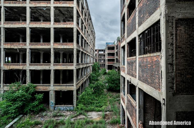 Looking down an alley between the brick and concrete shells of abandoned factory buildings. The skies are overcast and the ground is overgrown