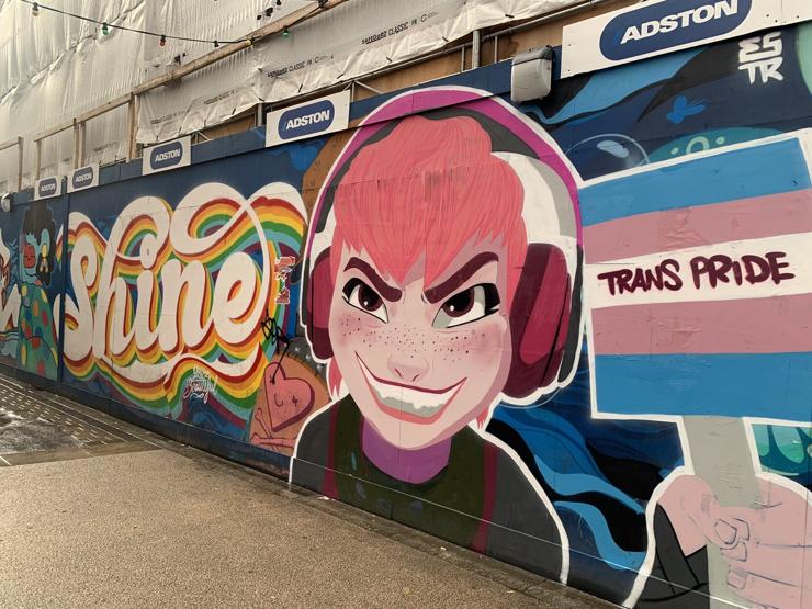 A mural wall depicting the film version of Nimona with a menacing toothy grin, wearing pink headphones. She is holding a square sign with the trans flag, and the words Trans Pride.

To the left is the word “Shine” with a rainbow colored shadow.