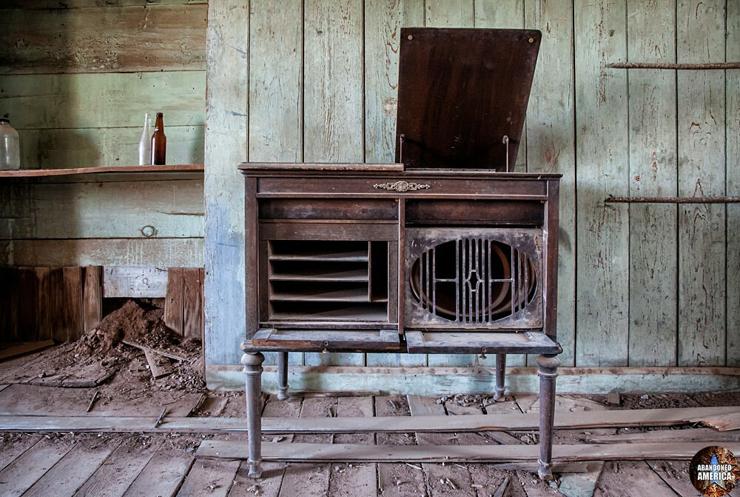 An antique console record player with its lid propped open. The walls and floors are wooden boards and to the left in a recession in the wall there is a rough wooden shelf with old bottles on it