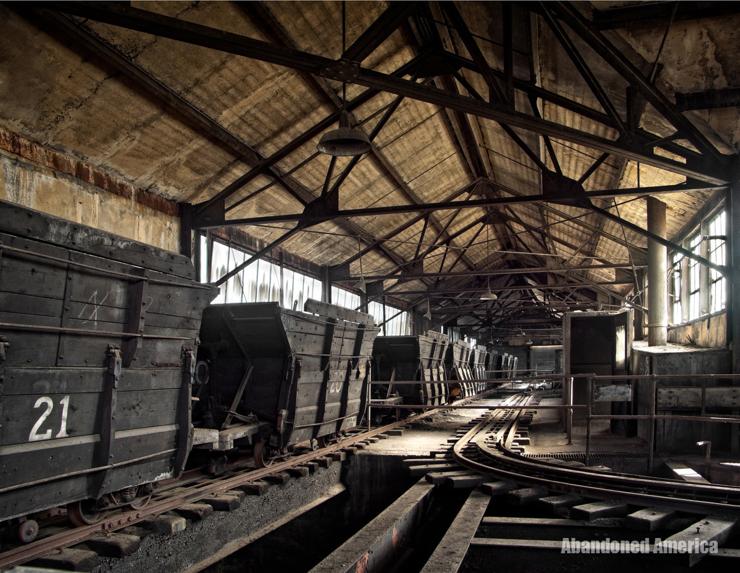 A concrete corridor lined with railroad tracks. Black wooden coal cars sit on the tracks, stretching into the distance. 