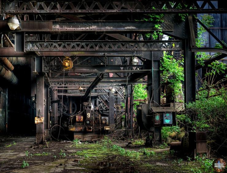 The underside of an overgrown steel trestle. In the distance a rusty black scale car with the number 9 painted on it in white rests beneath an overhead rail.