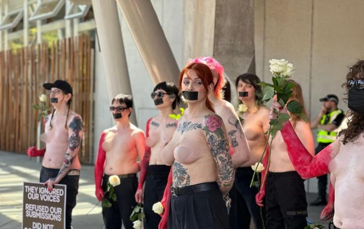 8 topless women with black masking tape over their mouths, in front of a building. A police officer is monitoring behind them. The photograph has been censored to obscure breasts and/ or nipples.