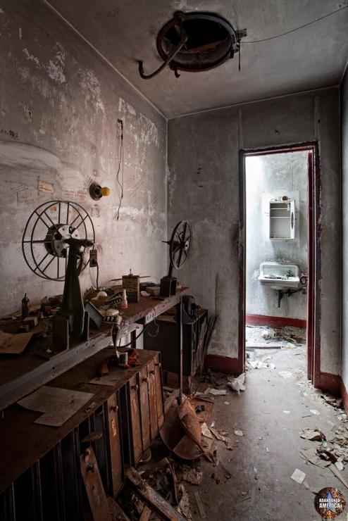 A bench in a darkened room with two reels for winding film and containers underneath for storing film containers. A door behind the bench is open into a bright but decayed bathroom, in which a sink and medicine cabinet are visible