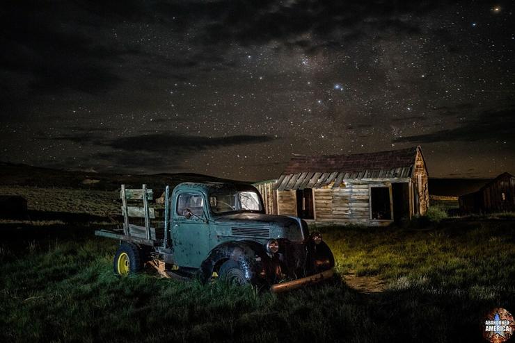 An antique pickup truck sitting in front of a wooden shed. Though a few clouds are overhead the stars are shining brightly.